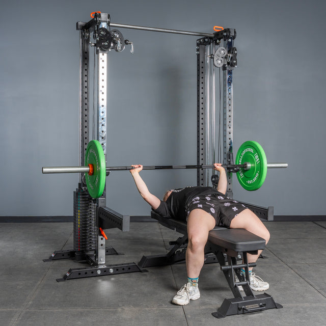 A person lies on a bench in a gym, performing a bench press with a barbell loaded with green plates, using Bells of Steel Cable Tower Squat Stands for support.