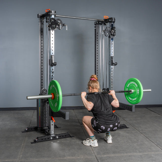 A person with light hair wearing black workout clothes and white shoes performs a squat with a barbell loaded with green plates on Bells of Steel Cable Tower Squat Stands in a gym with gray walls and floor.