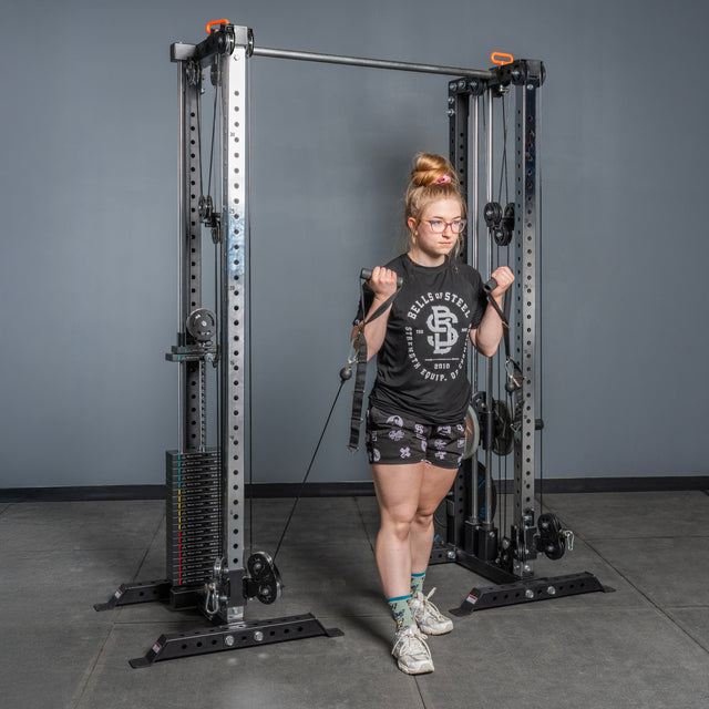 A woman in glasses and workout attire uses the Bells of Steel Cable Tower Squat Stands to perform a bicep curl exercise in a gym with gray walls and a concrete floor.