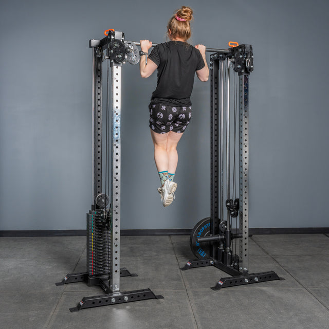 A person with their hair in a bun performs a pull-up on the Bells of Steel Cable Tower Squat Stands in a gym with gray walls and a cement floor, wearing a black shirt, patterned shorts, and colorful socks.