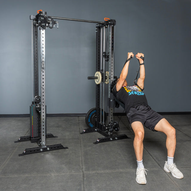 A person performs a cable chest press on the Bells of Steel Cable Tower Squat Stands, leaning back with arms extended and holding the handles, in a gym with gray walls and floor.