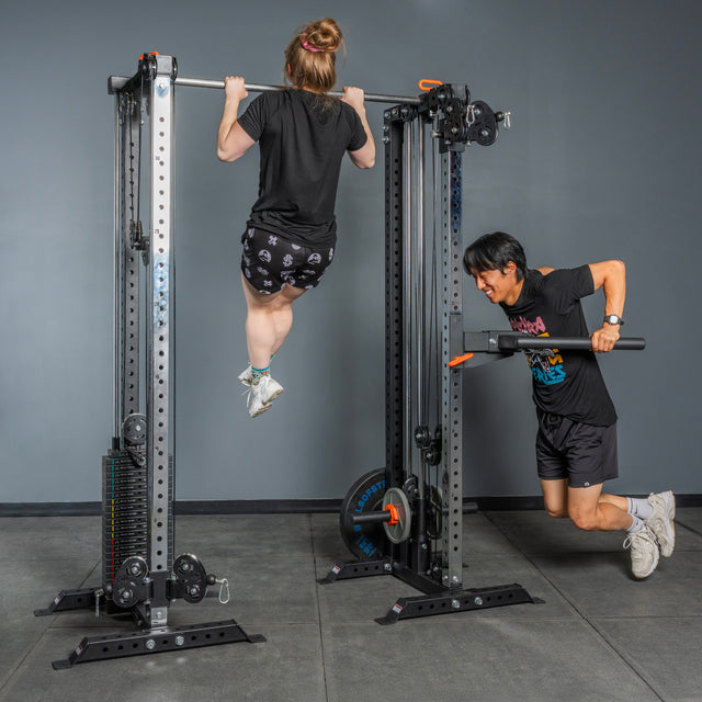 A woman does a pull-up while a man performs a triceps pushdown on the Bells of Steel Cable Tower Squat Stands in a gym with gray walls and floor, highlighting the versatility of this home gym equipment.