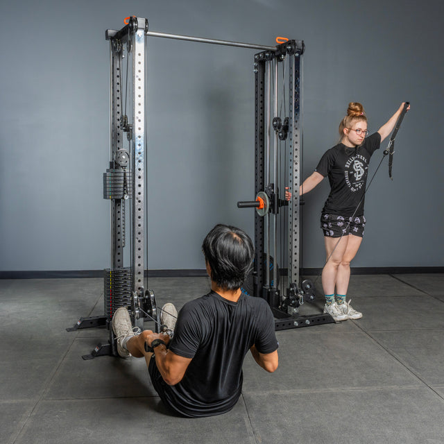 A woman uses the Bells of Steel Cable Tower Squat Stands for a single-arm lateral raise, while a man does a cable row seated on the floor. Both are in a gym with gray walls and flooring.