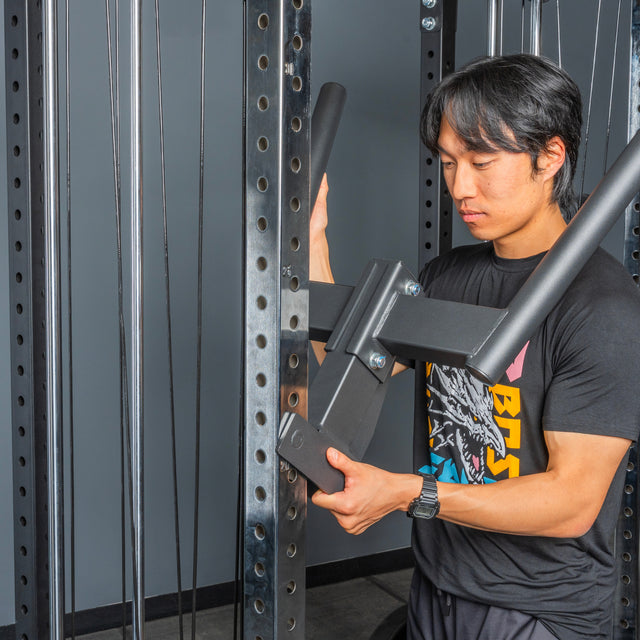 A person in a black graphic t-shirt adjusts a large metal attachment on the Bells of Steel Cable Tower Squat Stands, preparing equipment in a fitness setting.