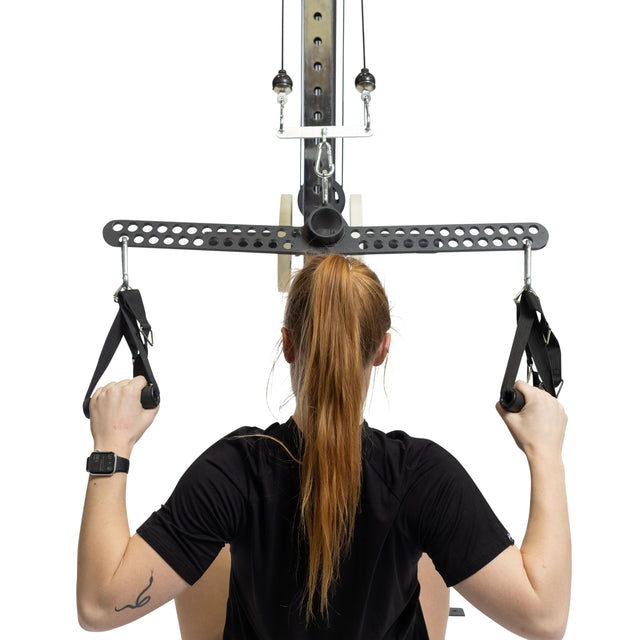 A woman with long red hair in a ponytail uses the Bells of Steel 30 D-Handle Bar Cable Attachment with Carabiners, gripping the adjustable nylon handles firmly as she pulls down. She wears a black shirt, smartwatch, and has a tattoo on her left forearm.