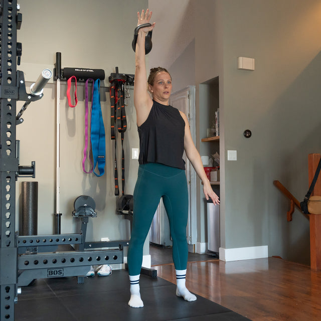 A woman in athletic wear lifts the Bells of Steel 6-12KG Adjustable Kettlebell overhead with one arm in a home gym, standing on a mat with other equipment and resistance bands visible in the background.