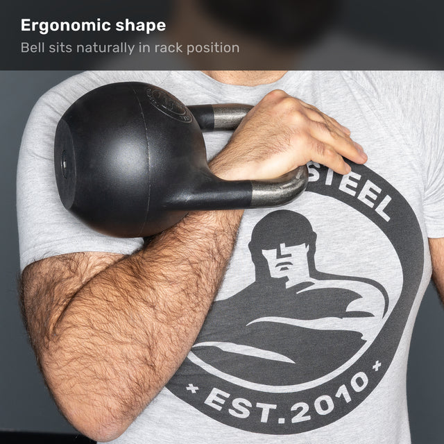 A person holds a Bells of Steel 6-12KG Adjustable Kettlebell, competition-style and black in color, in the rack position against their chest. They wear a gray t-shirt featuring a graphic of a muscular figure and the text âSTEEL EST. 2010.â.