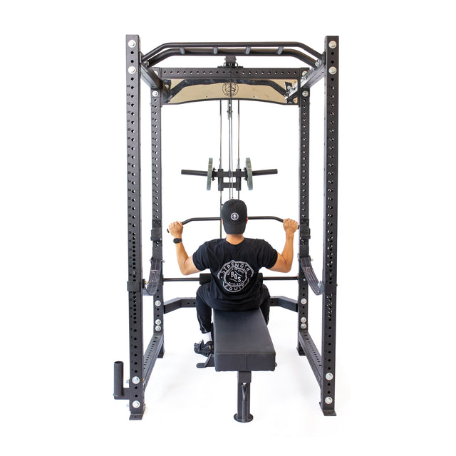 A person in a black T-shirt and cap uses the Bells of Steel Lat Pulldown & Low Row Rack Attachment – Hydra & Manticore for cable exercises inside a power rack, viewed from behind against a white background.