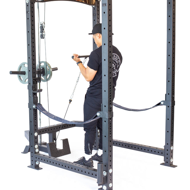 A person in black athletic wear uses the Bells of Steel Lat Pulldown & Low Row Rack Attachment – Hydra & Manticore, performing a seated row inside a power rack with Olympic weight plates loaded and safety straps attached.