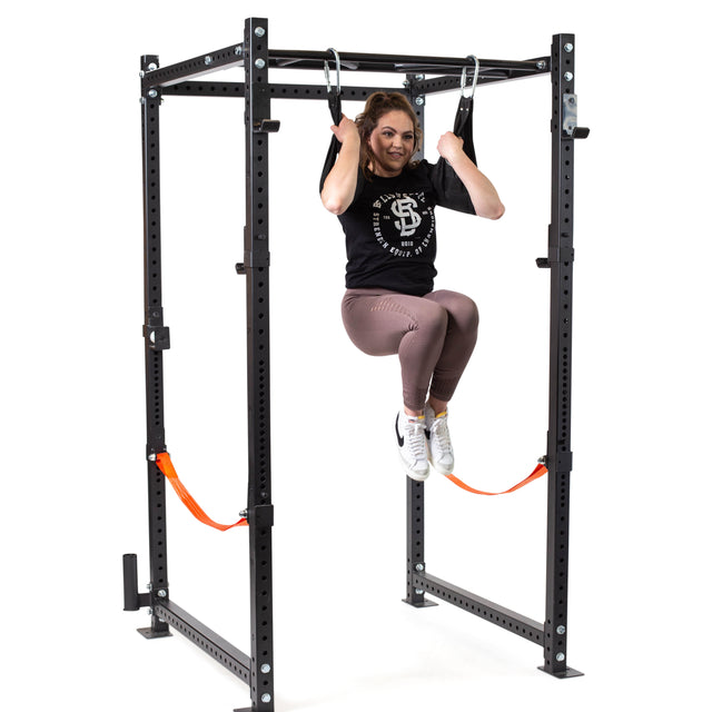 A woman uses Bells of Steel Hanging Ab Straps, suspended from a black metal workout rack, supporting her arms for a core workout with knees bent and feet off the ground, smiling at the camera in black gym attire.