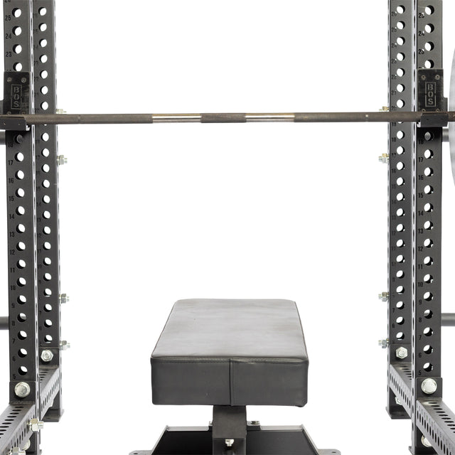 A flat weight bench sits under a racked barbell in a power rack outfitted with Bells of Steel Standard J-Cups, which have UHMW protection and multiple adjustment holes and markings.