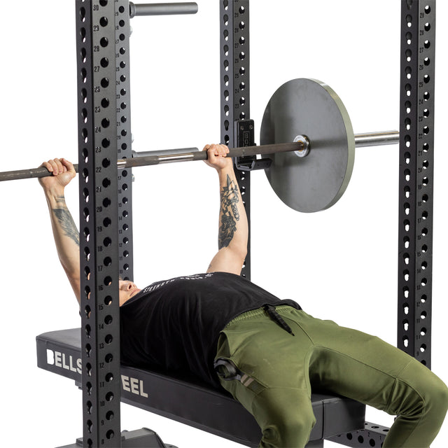 A person in green pants and a black shirt lies on a bench inside a power rack, gripping a barbell resting on Bells of Steel Standard J-Cups, preparing to perform a bench press.