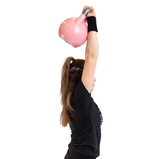 A woman wearing Bells of Steel’s BellGuard Kettlebell Wrist Guards (Pair) and a black T-shirt lifts a pink kettlebell overhead with one arm, viewed from the side against a white background.