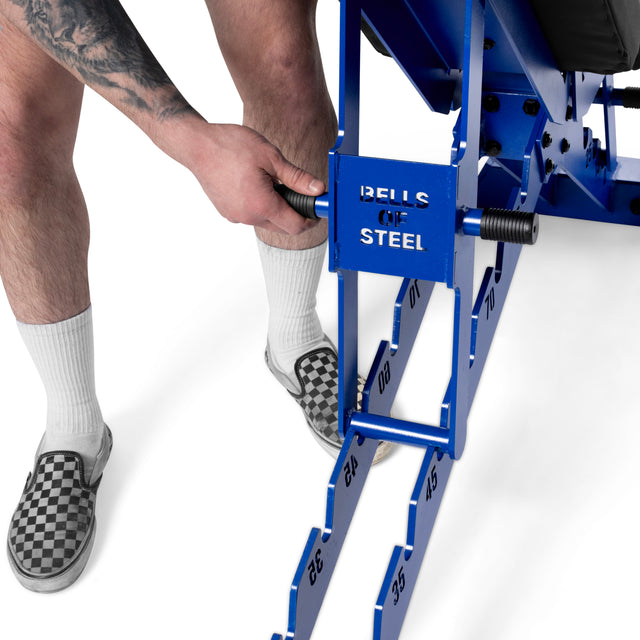 A person in checkered slip-on shoes and white socks adjusts a Bells of Steel Buzz-Saw Heavy-Duty Adjustable Bench, which features numbered incline settings for space-efficient storage.