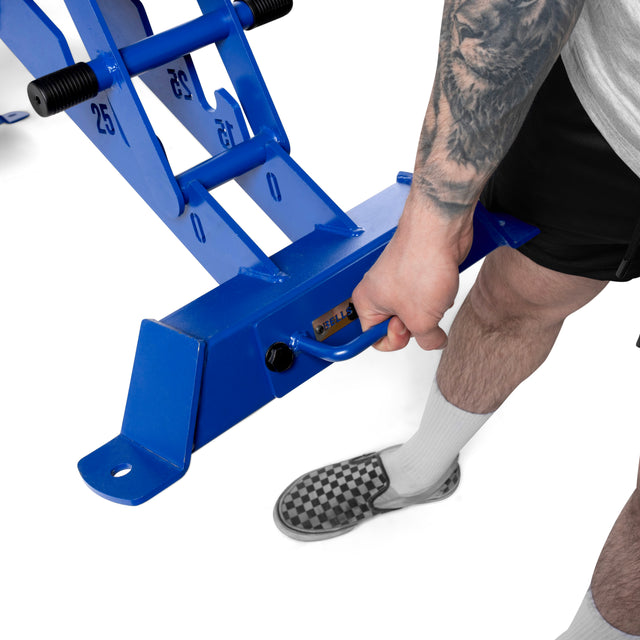 A tattooed individual in shorts, socks, and checkered slip-ons grips the blue handle of a Buzz-Saw Heavy-Duty Adjustable Bench by Bells of Steel, marked "25," against a simple white backdrop.