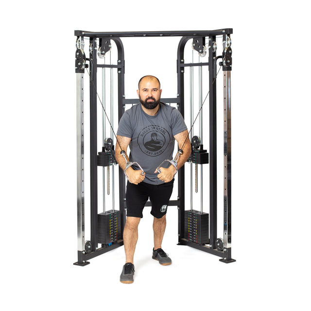 A man uses the Bells of Steel Functional Trainer - Weight Stack, gripping handles with both hands. He wears a gray t-shirt, black shorts, and athletic shoes, standing in a staggered stance against a white background.