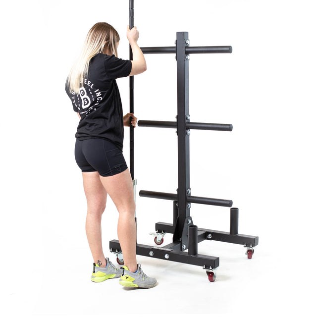 A woman with long blonde hair stands next to the Bells of Steel Bumper Plate Weight Tree And Bar Holder, holding a vertical pole attached to the storage rack on wheels.