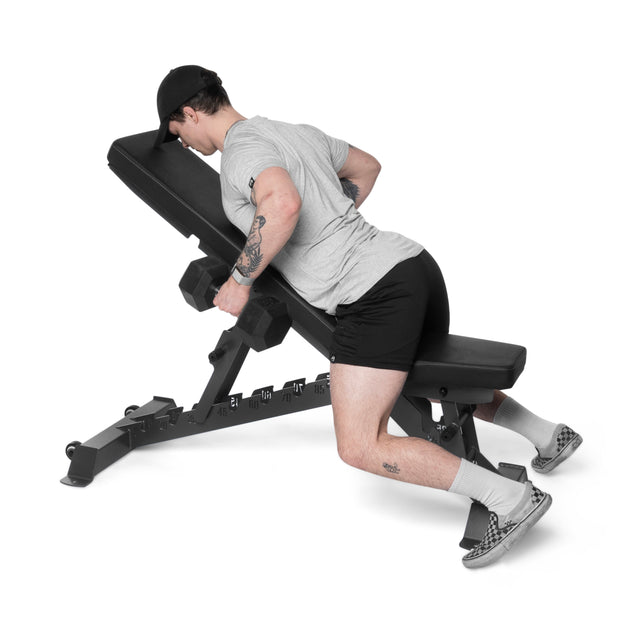 Kneeling on a Buzz-Saw Heavy-Duty Adjustable Bench from Bells of Steel, a person in a gray t-shirt, black shorts, white socks, and checkered sneakers lifts dumbbells, using the sturdy equipment for support while their head faces down.