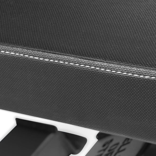 Close-up of the black textured seat with white stitching from the Buzz-Saw Heavy-Duty Adjustable Bench by Bells of Steel. The blurred background highlights the contrast of textures and lines.