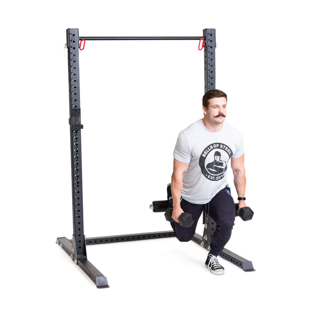 A man uses the Bells of Steel Split Squat Leg Roller Rack Attachment to perform Bulgarian split squats, holding a dumbbell in each hand. He's wearing a gray t-shirt, black pants, and sneakers.