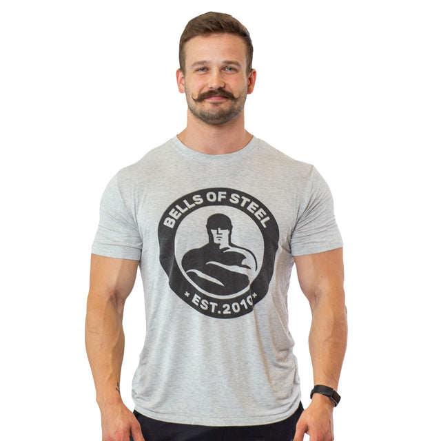 A smiling man with a mustache and short hair wears the Bells of Steel Bamboo T-Shirt, featuring sweat-wicking fabric and "Est. 2010" text, standing with arms relaxed at his sides against a white background.