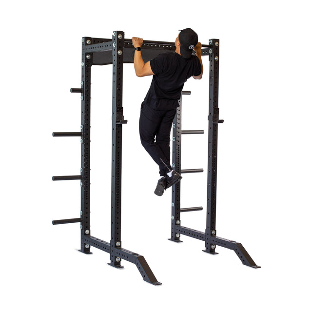 A person in black athletic wear and a cap does a pull-up on the Bells of Steel Hydra Collegiate Power Rack - Prebuilt (3" x 3", ⅝" Holes), featuring weight storage pegs, set against a white background.
