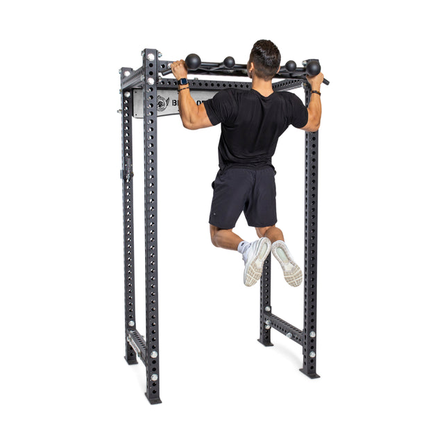 A person in a black shirt and shorts does a pull-up on the Bells of Steel Globe Pull Up Bar, mounted to a sturdy power rack, highlighting effective strength training, isolated on a white background.