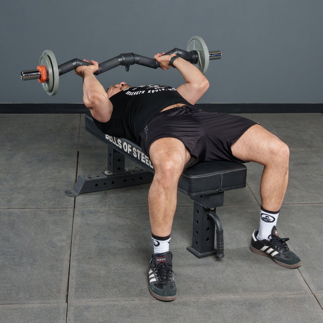 A person is bench pressing with the Bells of Steel Fat Bar - Olympic Curl Bar Cable Attachment in a gym, wearing a black shirt and shorts, white socks, and black athletic shoes to enhance grip strength.