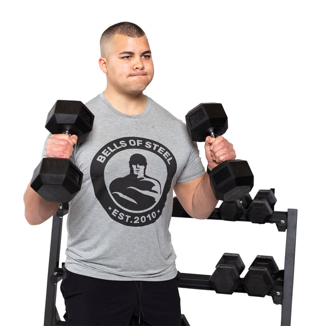 A focused individual wearing a gray Bells of Steel shirt lifts Ergo Rubber Hex Dumbbell Sets in front of a weight rack, captured mid-exercise against a white background.