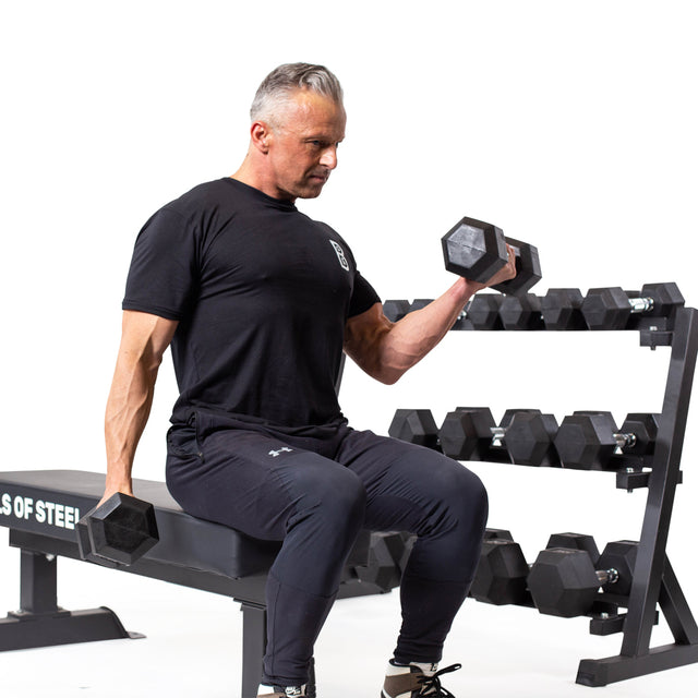 A man in black workout gear sits on a gym bench, performing bicep curls with Bells of Steel Ergo Rubber Hex Dumbbell Sets. A rack of these commercial-grade dumbbells is placed beside him against a white background.