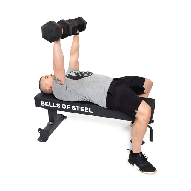 A person on a flat bench labeled "Bells of Steel" performs a dumbbell bench press with both arms extended, using Bells of Steel Ergo Rubber Hex Dumbbell Sets while wearing a gray t-shirt, black shorts, and black sneakers.