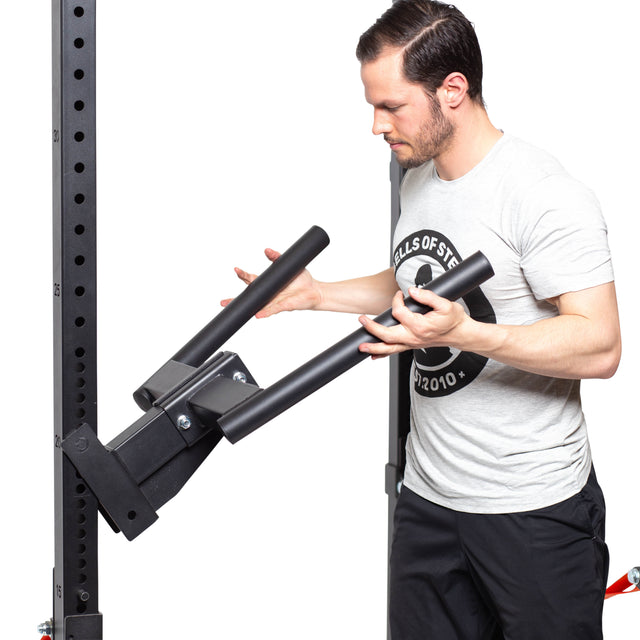 A man in a light gray t-shirt and black shorts adjusts a pair of angled metal bars on a Bells of Steel heavy-duty fitness rack, focused on positioning the Y Dip Bar Rack Attachment, an essential exercise accessory.