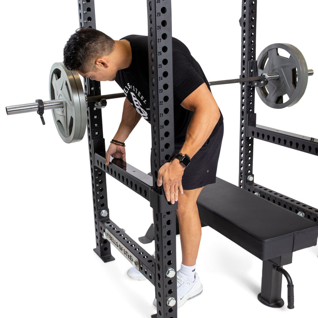 A person in black adjusts Bells of Steel Flip-Down Safeties on a power rack, ensuring training safety as a barbell with weight plates is set over a bench for bench press.