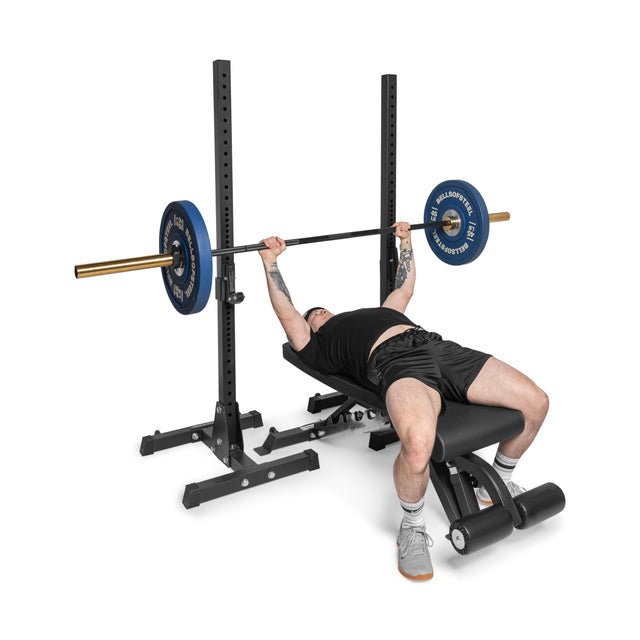 Wearing a black shirt, shorts, gray socks, and showcasing arm tattoos, a person uses the Bells of Steel Flat/Incline/Decline Weight Bench - Commercial to perform a bench press with precision on a barbell loaded with blue weights.