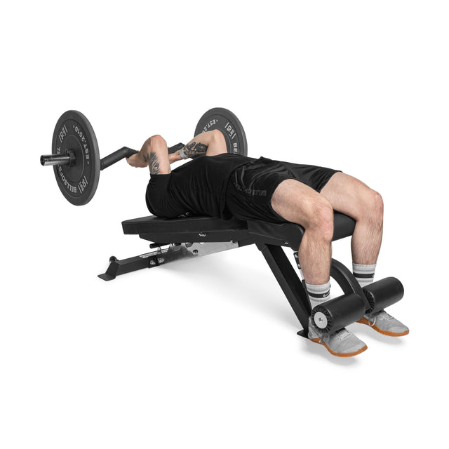 A person on a Bells of Steel Flat/Incline/Decline Weight Bench - Commercial performs a bench press with a barbell. Wearing a black shirt, black shorts, and gray shoes, the versatile bench aids in optimal muscle targeting while their feet secure under the metal roller.