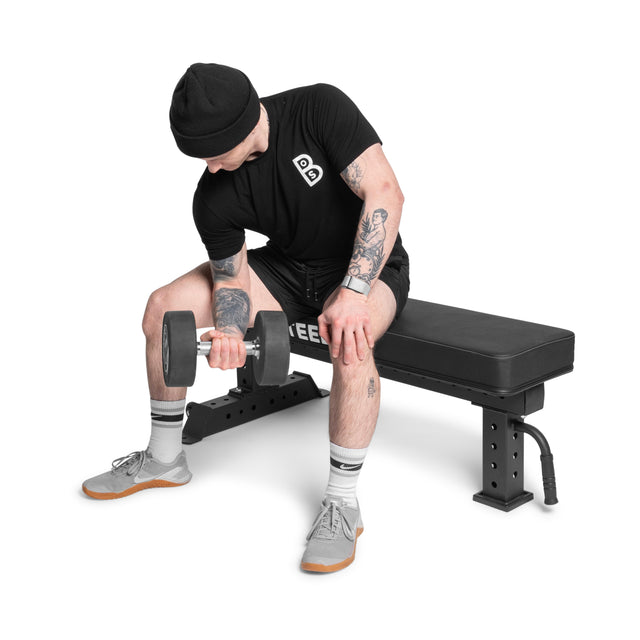 Dressed in black with a beanie, gray socks, and shoes, a person focuses on a dumbbell curl with their right arm while seated on the Bells of Steel Powerlifting Flat Bench. The bench's premium vinyl finish enhances their intense workout against the stark white background.