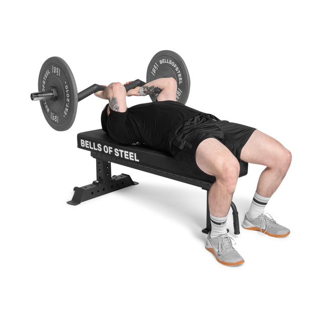 Dressed in black shorts and a T-shirt, an individual performs a barbell bench press on the Bells of Steel Powerlifting Flat Bench. With precision and ease, they lift the barbell while lying flat against a white backdrop, wearing socks and athletic shoes.
