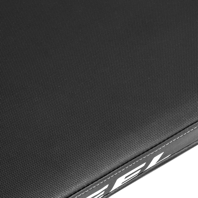Close-up of a textured dark gray surface with visible stitching on the edge, typical of the Bells of Steel Powerlifting Flat Bench. A partial view of a white printed design appears on the side over a sleek black background.