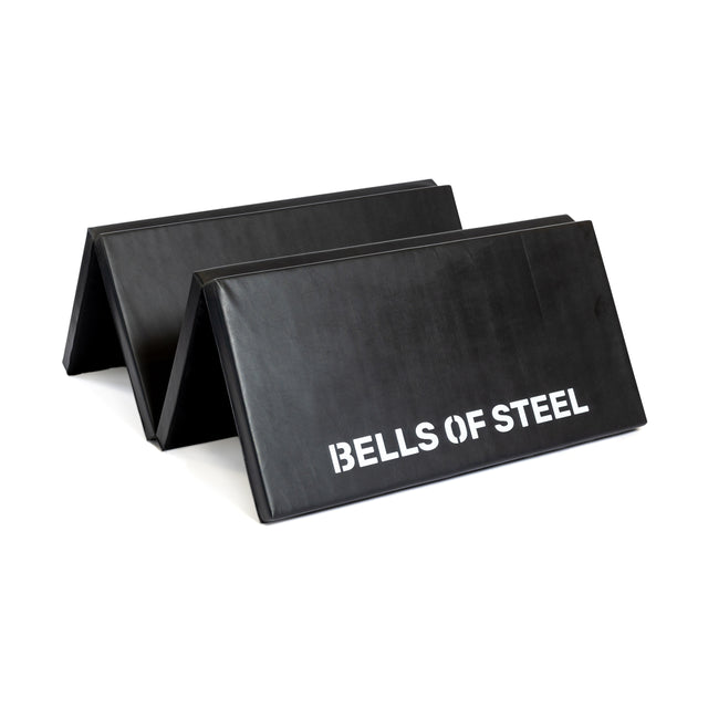 Two Four Fold Mats by Bells of Steel, made with plush foam for comfort and “BELLS OF STEEL” in white text, stand zigzagged on a white background.