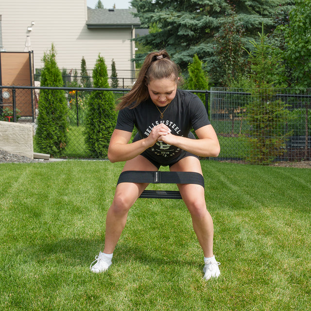 A woman demonstrates the Bells of Steel Ultimate Backyard Training Bundle, performing squats with a resistance band secured around her thighs. She trains outdoors on a grassy lawn, dressed in a black t-shirt, black shorts, and white sneakers. The backyard gym scene is complete with shrubs, a fence, and houses in the background.