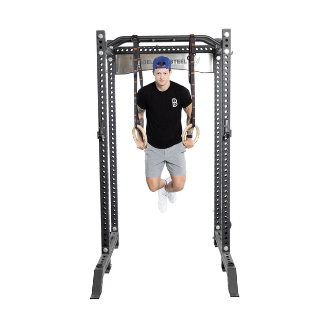 A person in a blue cap, black shirt, and gray shorts is doing a core workout using Bells of Steel Gymnastics Rings on a rack, suspended for bodyweight training while holding themselves up with slightly bent legs.