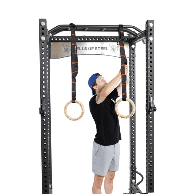 Wearing a blue cap, black t-shirt, and gray shorts, a person prepares for an intense core workout with Bells of Steel Gymnastics Rings. They hang the rings from adjustable straps on a black metal power rack featuring the "BELLS OF STEEL" banner at the top.