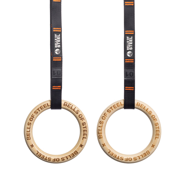 Bells of Steel's wooden Gymnastics Rings, with "BELLS OF STEEL" engraved, hang from black and orange straps featuring the logo. The setup is perfect for a core workout and stands out against the white background.