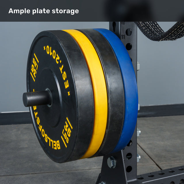 Close-up of weight plates in black, blue, and yellow neatly stacked on a storage rack, set against the gray walls and floor of a gym environment. The scene highlights the ideal setup for a home gym or the Bells of Steel Manticore Half Rack - Prebuilt (3" x 3", 1" Holes) arrangement.