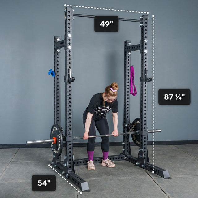 A person prepares to lift a barbell inside the Bells of Steel Manticore Half Rack - Prebuilt (3" x 3", 1" Holes), which features robust 3x3 uprights. The rack’s dimensions are clearly labeled: 49 inches wide, 54 inches deep, and standing at 87 ¼ inches tall—making it an ideal choice for any serious home gym setup. Purple and blue bands hang on the rack.