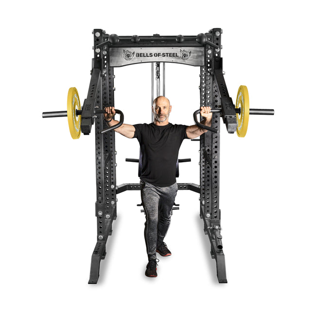 A man performs an exercise with one leg forward, holding the Bells of Steel Lever Arms Rack Attachment at shoulder height on a power rack with Olympic weight plates, against a white background.