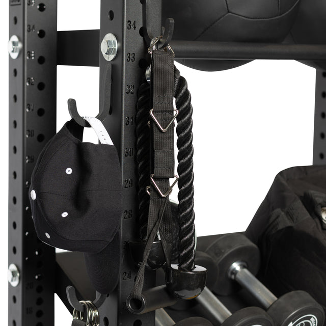 A Bells of Steel Utility Hook displays a black baseball cap, tricep rope attachment, and black medicine ball on a gym rack. Dumbbells and a gym bag rest below, highlighting an efficient gym storage solution.