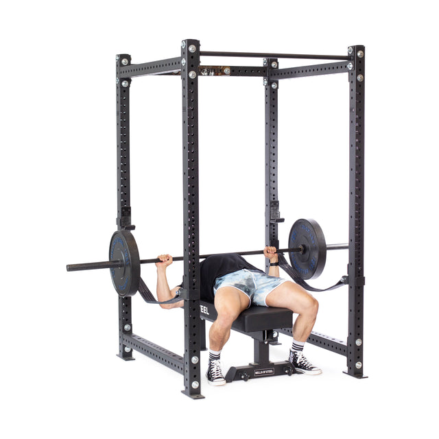 Wearing a black shirt, patterned shorts, and sneakers, a person bench presses with weight plates on a Hydra Four Post Power Rack - Prebuilt (3" x 3", ⅝" Holes) by Bells of Steel, highlighting an intense home gym workout.