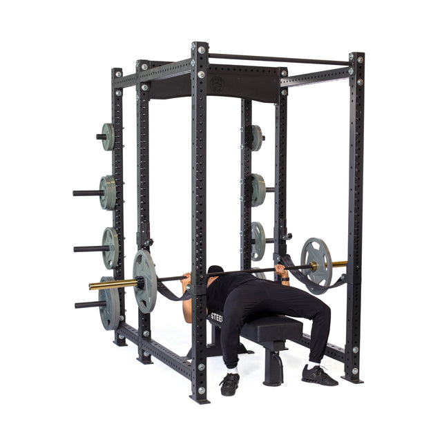 A person uses the Bells of Steel Hydra Six Post Power Rack (Prebuilt, 3" x 3", ⅝" Holes) for bench presses, taking advantage of built-in side peg weight storage—making it a top choice for home gyms.