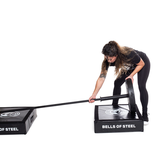 A woman prepares for landmine exercises by placing one end of a barbell into a Bells of Steel base and using Deadlift Pads (Pair) for protection. She wears a black shirt and leggings while gripping the other end.